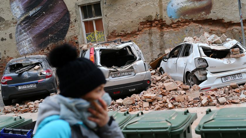 Uma croata com máscara passa diante de carros destruídos por escombros após o sismo de Zagreb. Foto: Reuters